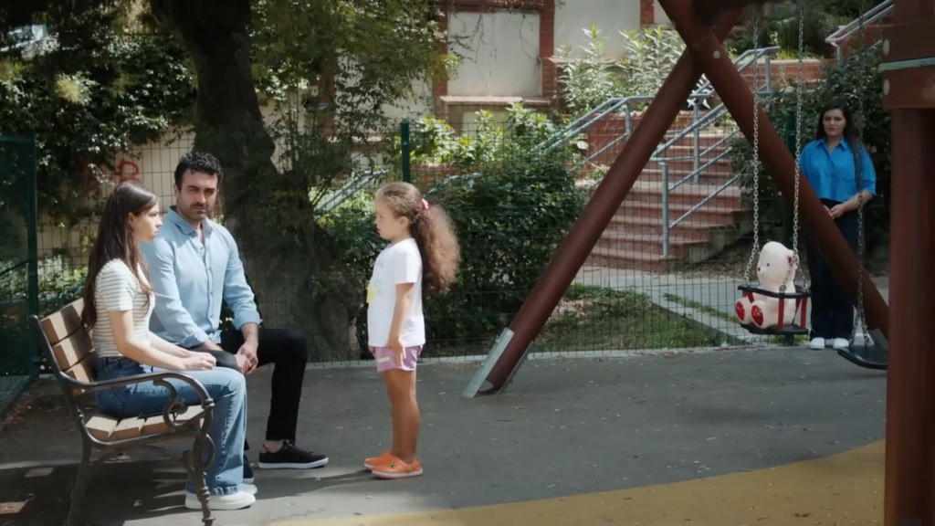 Ayse i Ferit siedzą na ławce w parku, a Doga stoi przed nimi.