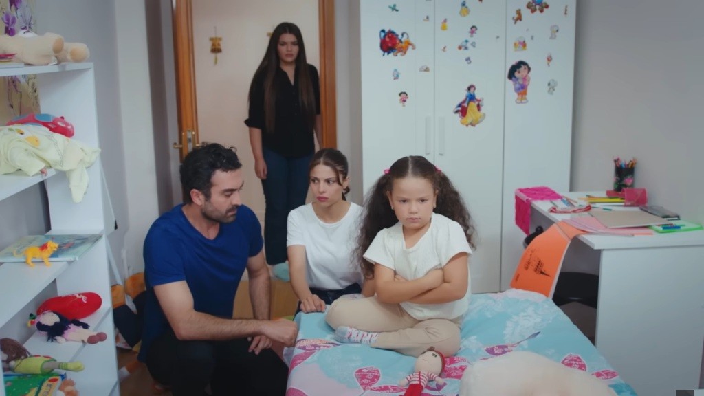 Ayse i Ferit próbują porozmawiać z córką, ale ona siedzi na łóżku z założonymi rękami i odwrócona do nich plecami.