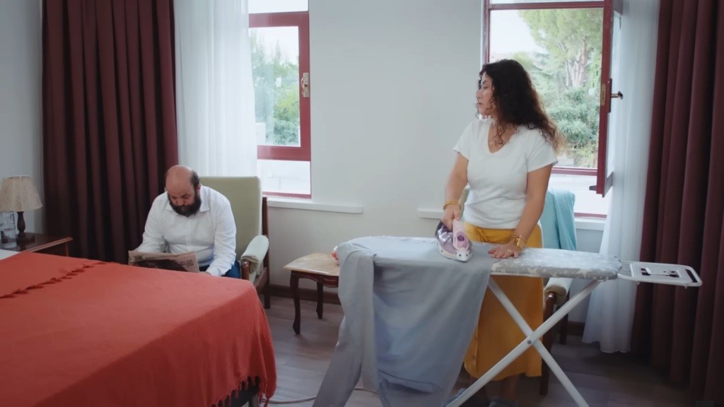 Cansel prasuje ubrania i patrzy na czytającego gazetę męża.