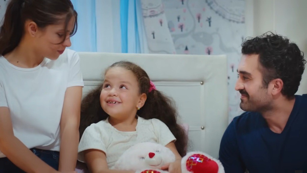 Doga siedzi na łóżku i uśmiecha się. Obok znajdują się jej rodzice.