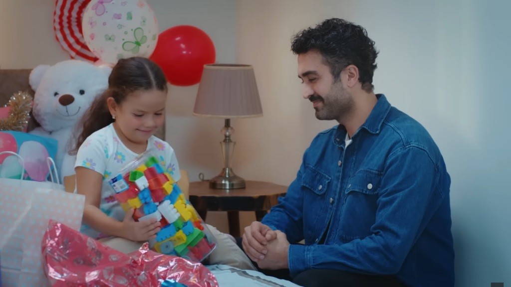 Doga trzyma w rękach opakowanie z klockami. Obok kuca Ferit. Oboje są szczęśliwi.