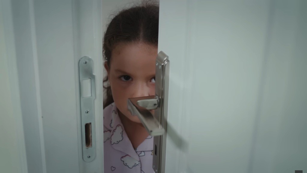 Doga stoi przy uchylonych drzwiach pokoju, skąd podsłuchuje rozmowę rodziców.