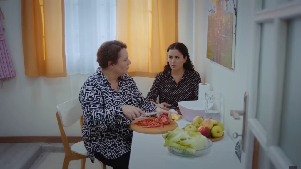 Adalet i Aynur siedzą przy stole w kuchni. Adalet kroi pomidora.