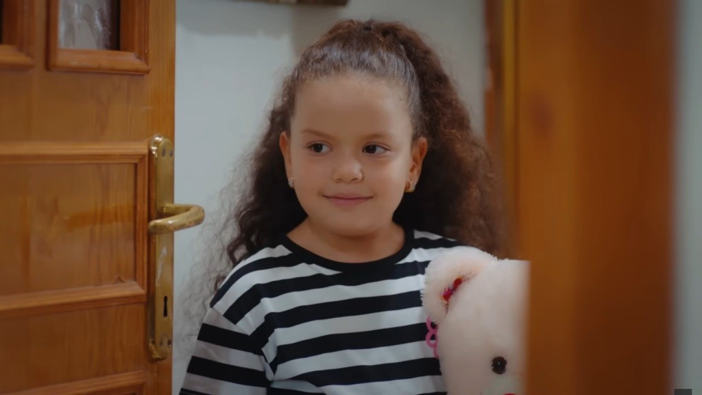 Doga trzyma w ręku misia i wygląda przez drzwi z delikatnym uśmiechem.