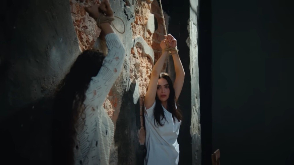 Nazli i Nana stoją przy ścianie ze związanymi i uniesionymi w górę rękami.