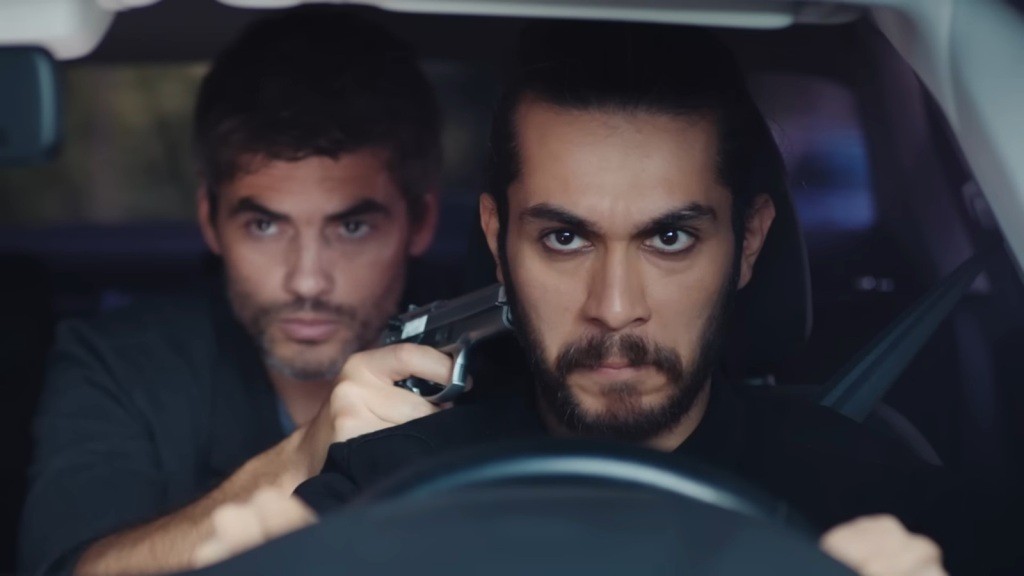 Wilka siedzi za kierownicą. Poyraz znajduje się za jego plecami i trzyma pistolet przy jego karku.