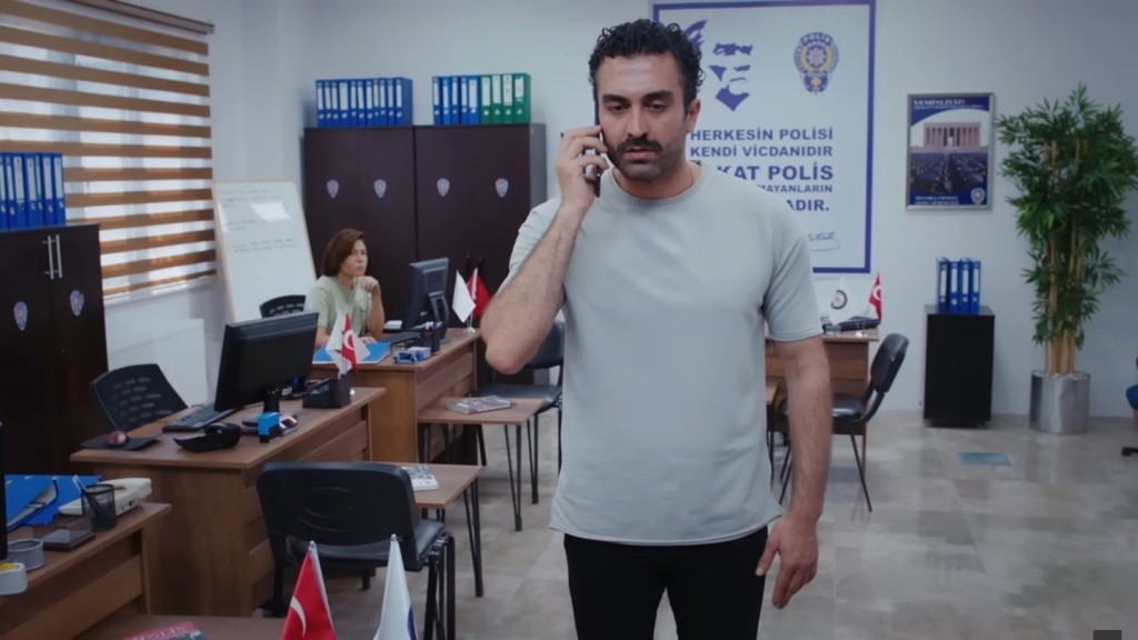 Ferit trzyma telefon przy uchu. Jego twarz wyraża zaniepokojenie. W tle Kara siedzi przy swoim biurku.