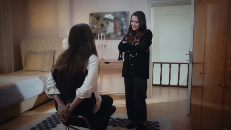 Derya stoi z założonymi na piersi rękami, patrząc na swoją siostrę, która jest przywiązana do krzesła.