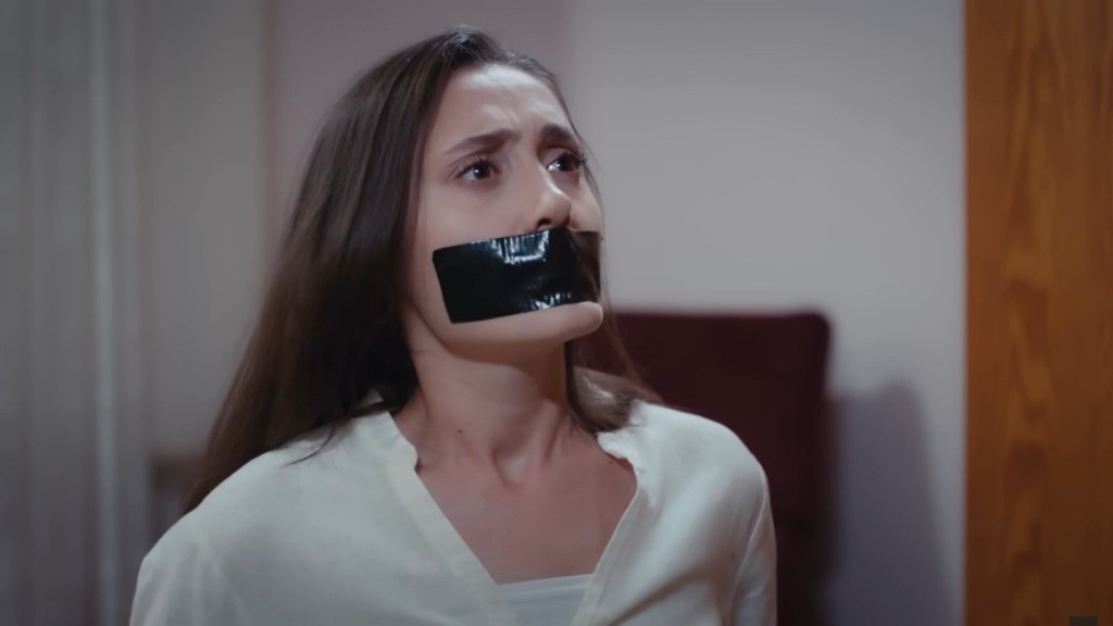 Zbliżenie na zakneblowaną Leylę, w której oczach widać wyraźny strach.