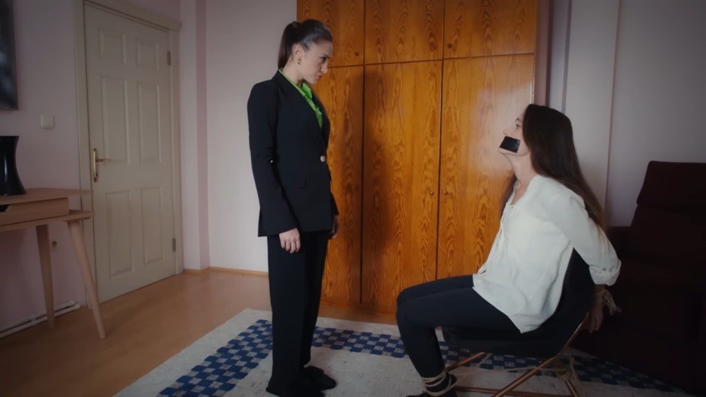 Ubrana w elegancki strój Derya stoi przed przywiązaną do krzesła Leylą.