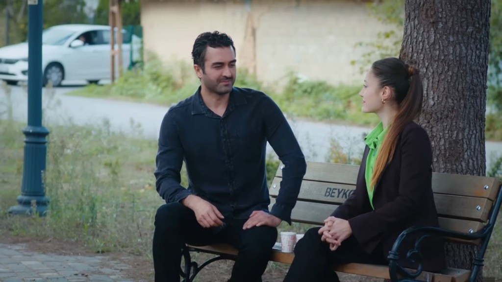 Ferit i Derya siedzą na ławce w parku. W tle, na drodze, widać zaparkowany biały samochód.