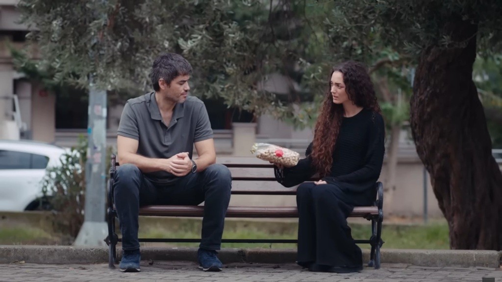 Poyraz i Nana siedzą na ławce. Dziewczyna wyciąga w jego stronę paczkę fistaszków.