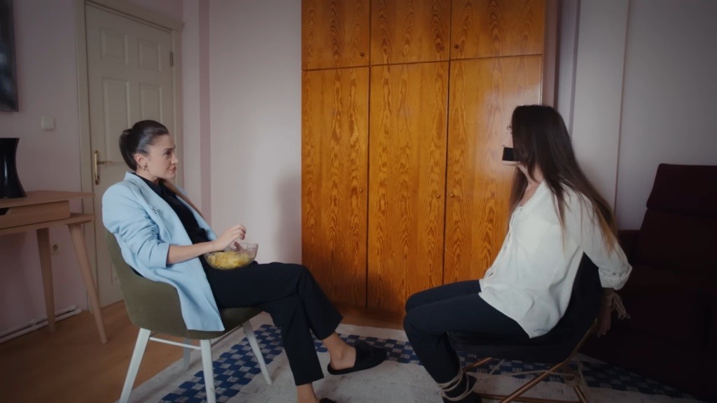 Derya siedzi przed związaną i zakneblowaną siostrą. Na kolanach trzyma szklanką miskę z czipsami.