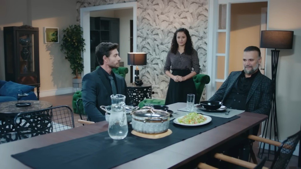 Prokurator i jego gość siedzą przy stole. Obok stoi Aynur.