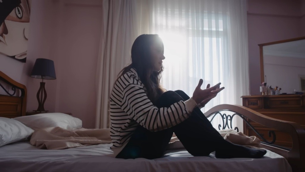 Derya siedzi na łóżku, wyciągając przed siebie ręce.