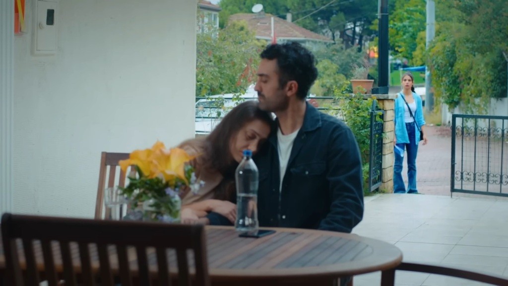 Derya opiera głowę na ramieniu Ferita, siedząc z nim przy stoliku na zewnątrz, podczas gdy Ayşe obserwuje ich z ukrycia zza bramy.