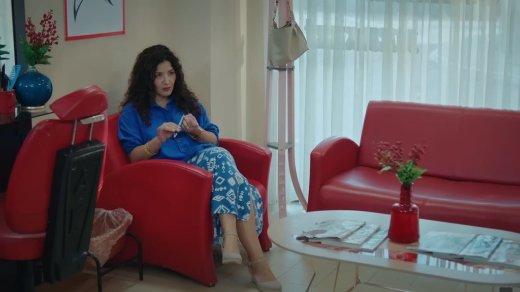 Cansel siedzi w fotelu i piłuje paznokcie, obserwując pracę Nany.