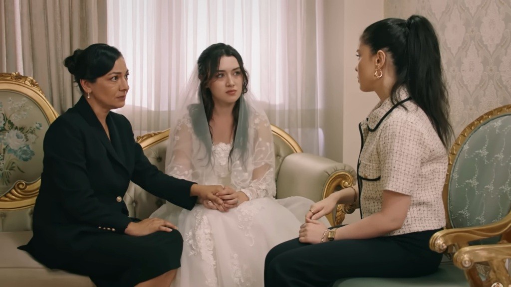 Zeynep ma na sobie suknię ślubną. Trzyma rękę matki. Obok siedzi Merve.