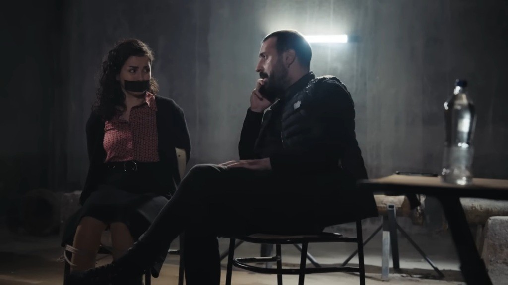 Związana i zakneblowana Aynur spogląda z przerażeniem na swojego porywacza, który rozmawia przez telefon.