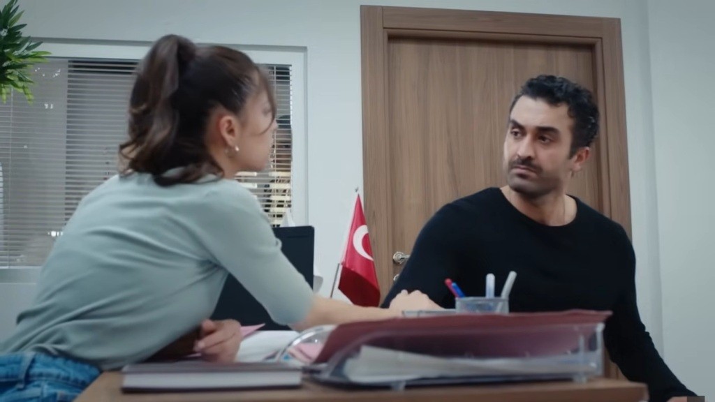 Ayse chwyta Ferita za rękę, siedząc naprzeciwko niego przy biurku.