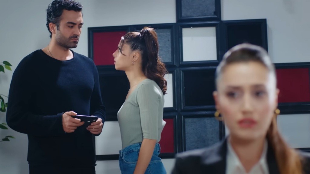Ayse i Ferit rozmawiają. Mężczyzna trzyma w rękach telefon. Na pierwszy planie widać rozmytą twarz Deryi, która sprawia wrażenie zaniepokojonej.