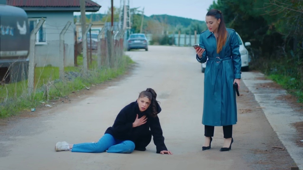 Ayşe leży na drodze, trzymając się za pierś, a obok niej stoi Derya, z telefonem w ręku, gotowa wezwać pomoc.