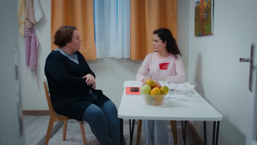 Adalet rozmawia z Aynur, siedząc przy stoliku.