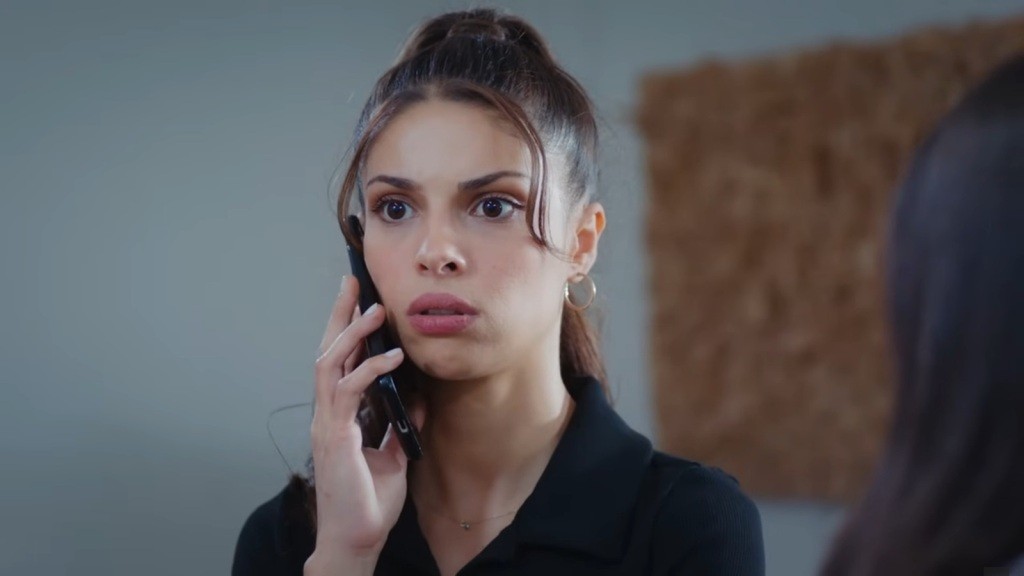 Ayse jest wstrząśnięta, trzymając telefon przy twarzy.