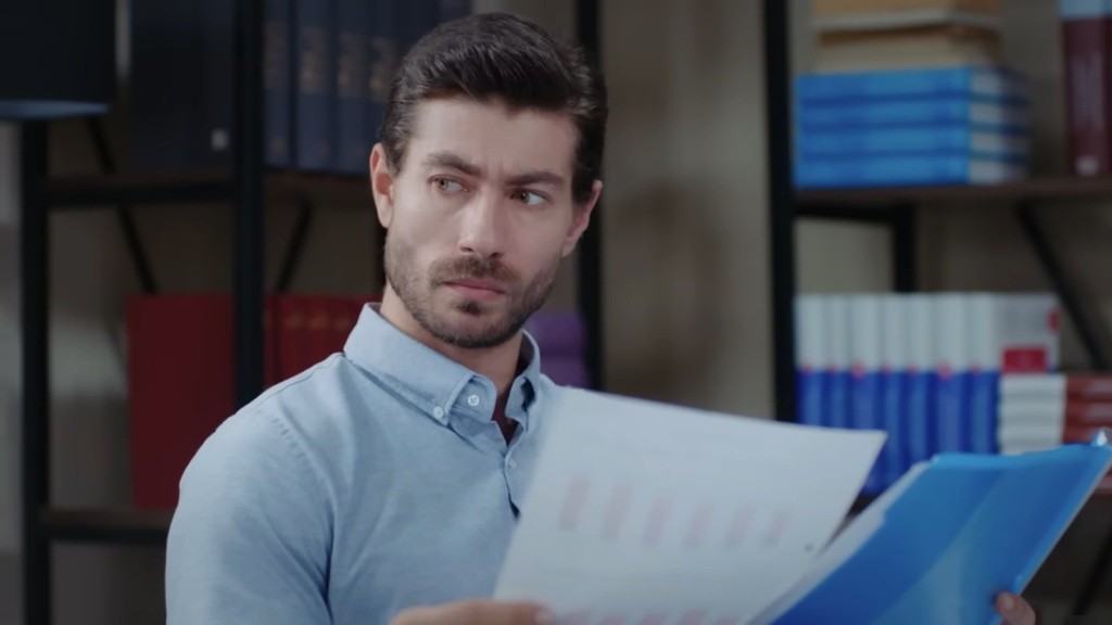 Prokurator Sinan trzyma dokument w ręku i zerka w bok.