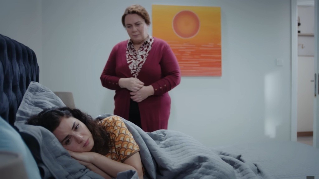 Aynur przytula głowę do poduszki. Przy jej łóżku stoi Adalet.