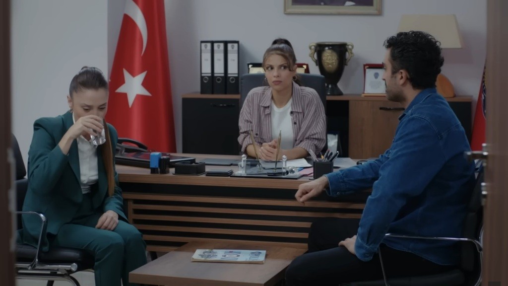 Derya pije wodę. Siedząca za biurkiem Ayse spogląda na Ferita.