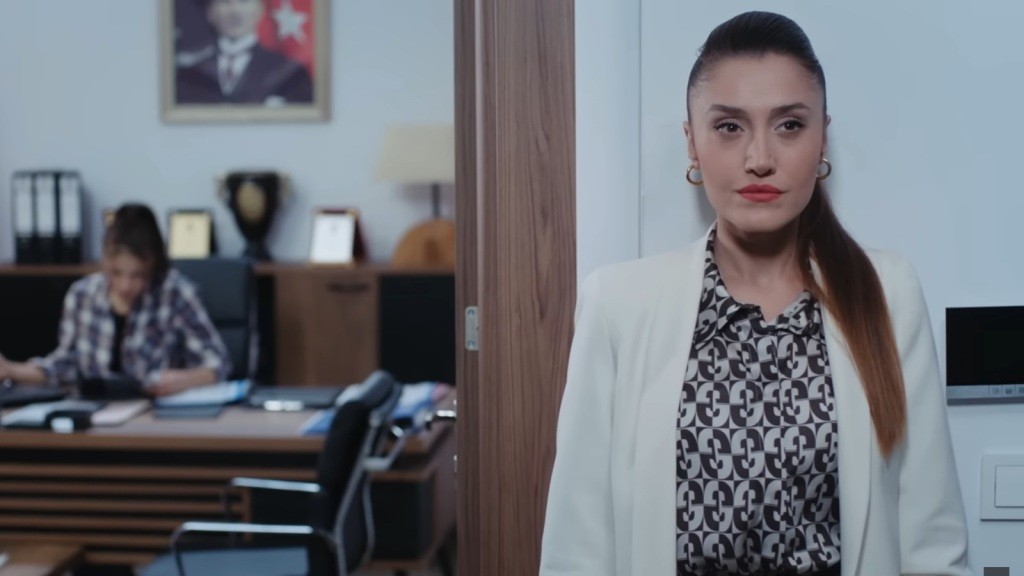 Derya stoi przy wejściu do gabinetu Ayse, opierając się plecami o ścianę.