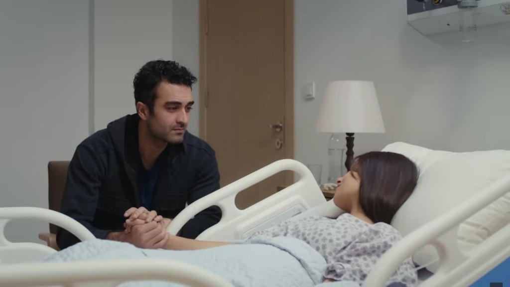 Ferit siedzi przy łóżku Ayse, delikatnie trzymając jej dłoń w swoich rękach.