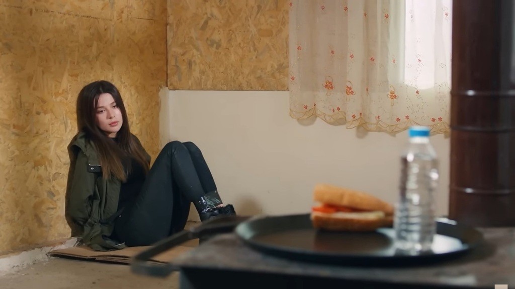 Związana Zeynep siedzi w kącie pokoju, patrząc na tacę z kanapką i butelką wody.