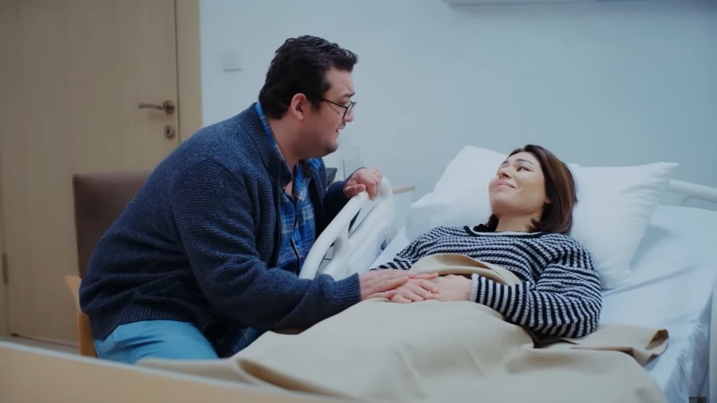 Ibrahim siedzi przy łóżku szpitalnym Kary. Dotyka jej dłoni. Oboje są uśmiechnięci.