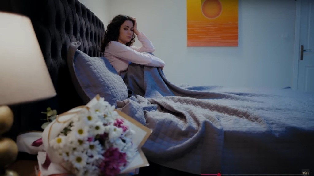 Aynur siedzi na łóżku, spoglądając na bukiet na szafce nocnej.