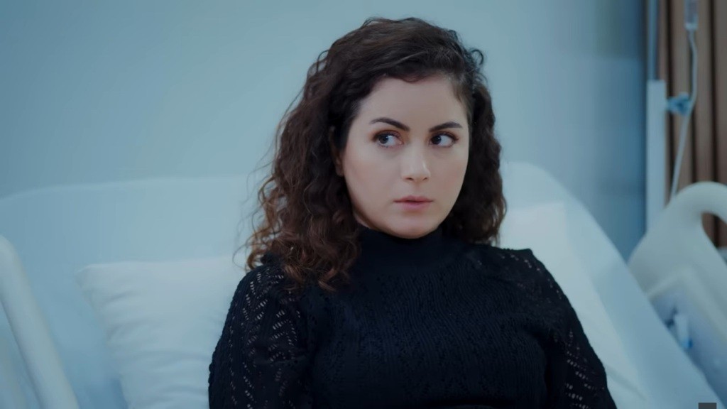 Aynur leży w pozycji półleżącej na szpitalnym łóżku.