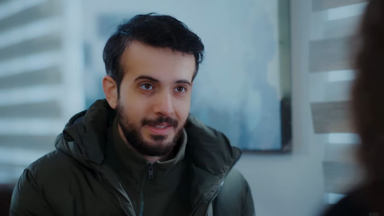 Mert siedzi naprzeciwko Nany i patrzy jej w oczy z lekkim uśmiechem.