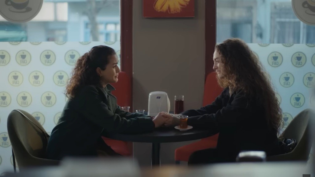 Aynur i Adalet siedzą przy stoliku, trzymając się za ręce.