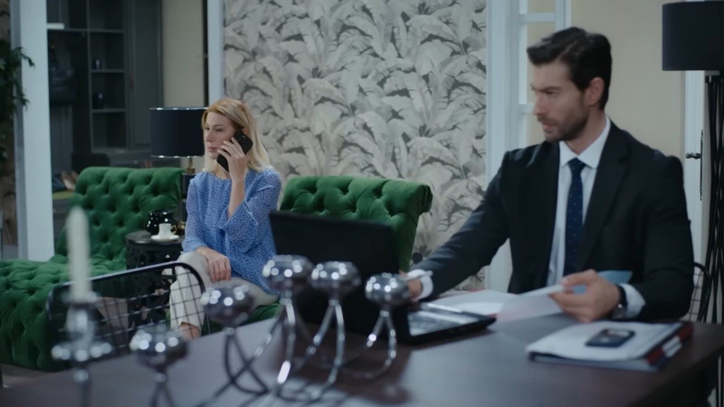 Pracujący przy stole prokurator przysłuchuje się rozmowie telefonicznej swojej matki, która siedzi w fotelu.