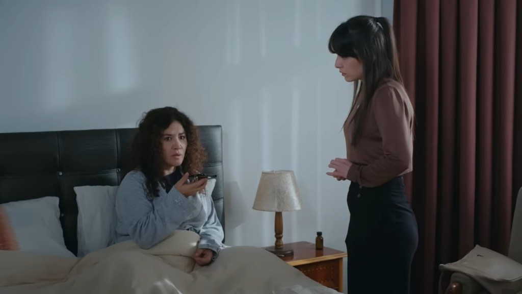 Cansel leży w łóżku i trzyma w ręku telefon. Sprawia wrażenie wystraszonej. Obok stoi Sibel.
