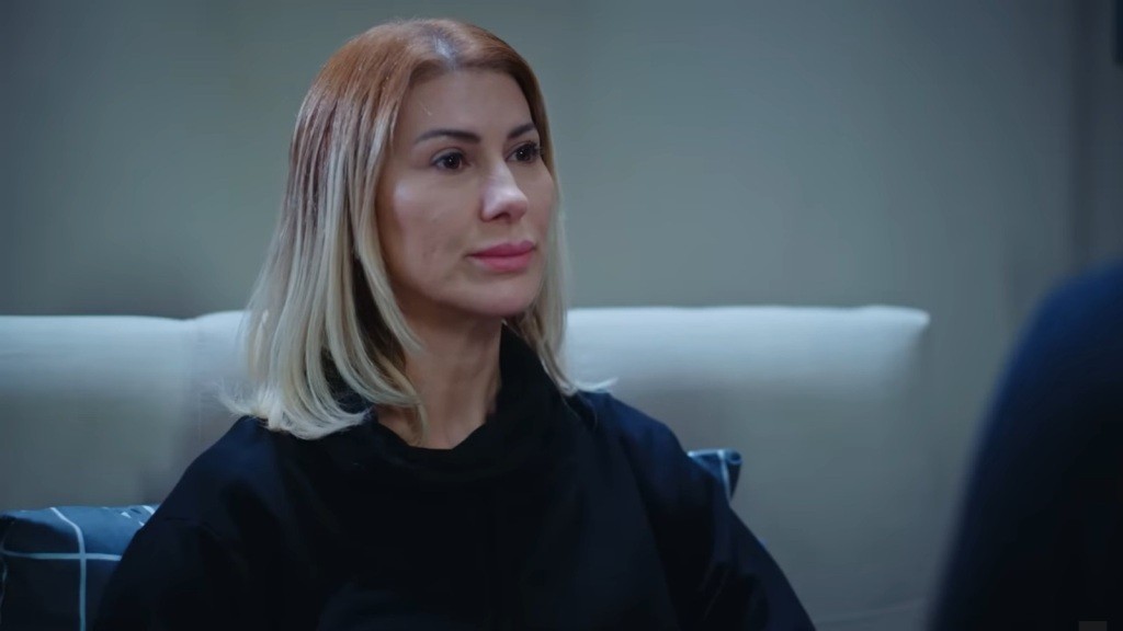 Isil siedzi oparta o wezgłowie łóżka.