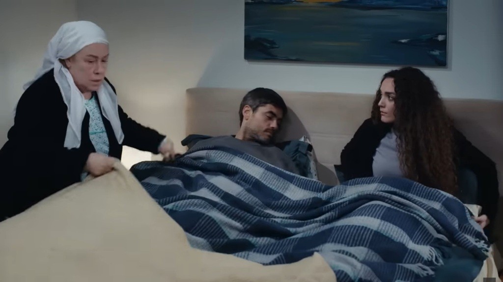 Poyraz i Nana leżą w jednym łóżku. Cennet poprawia kołdrę.
