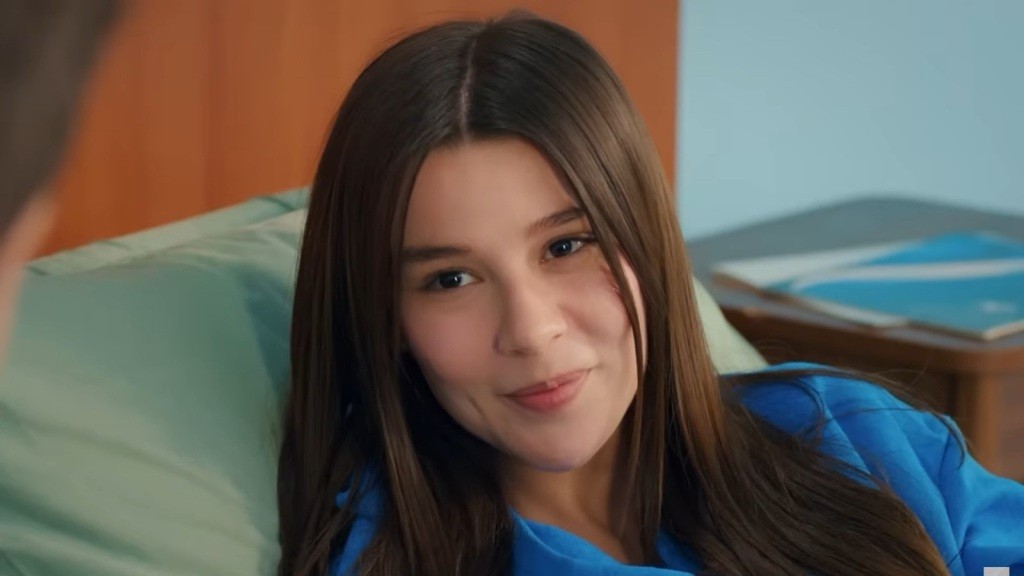 Zeynep leży na szpitalnym łóżku, uśmiechając się i unosząc głowę.