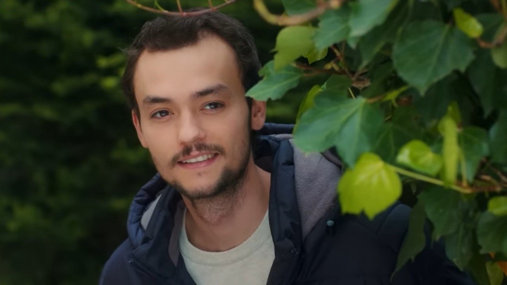Alper uśmiecha się, wyglądając zza zarośli.