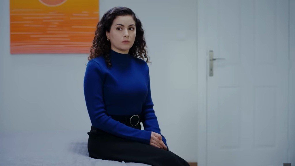 Aynur siedzi na łóżku i patrzy smutno.