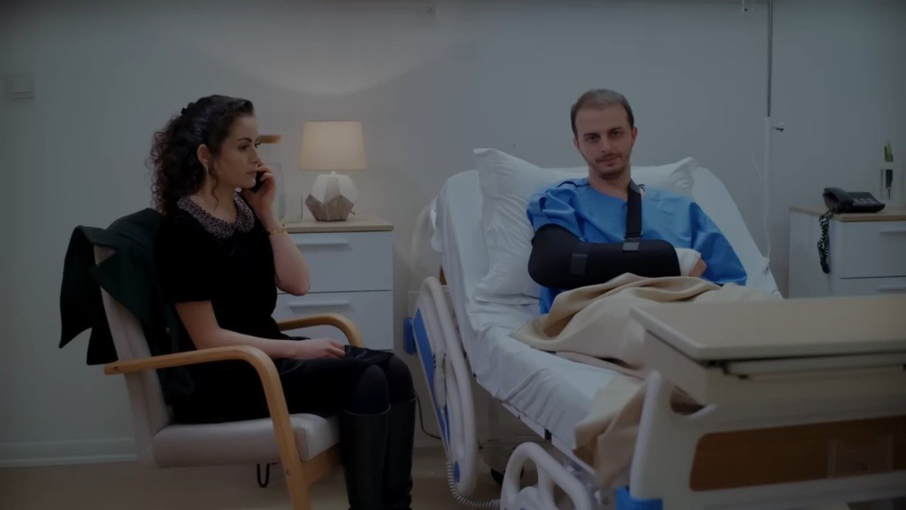 Kivanc leży w szpitalnym łóżku, z ręką unieruchomioną w temblaku. Obok niego siedzi Aynur, rozmawiając przez telefon