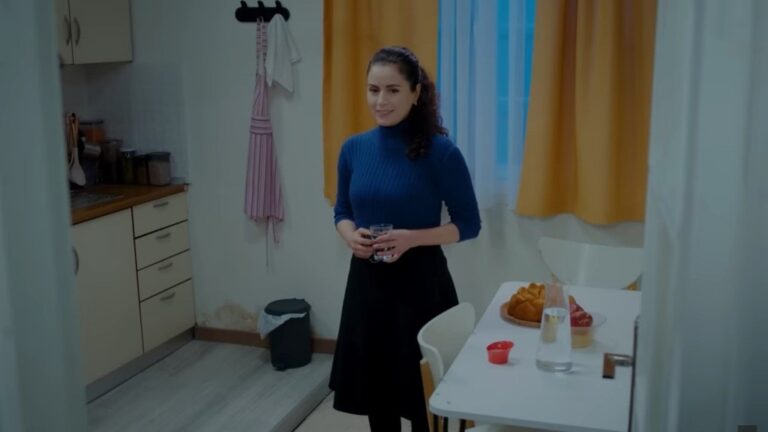 Aynur uśmiecha się, stojąc w kuchni ze szklanki wody w rękach.