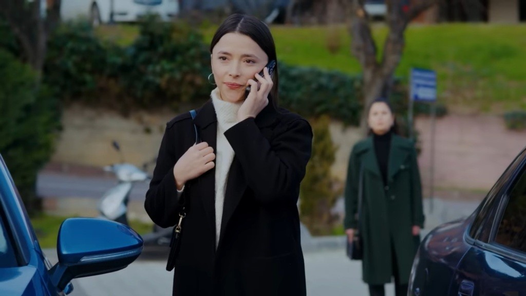 Ezgi rozmawia przez telefon na parkingu. Aynur stoi za jej plecami.
