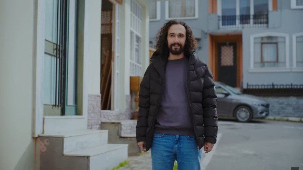 Semih stoi na ulicy przed warsztatem. Uśmiecha się przebiegle.